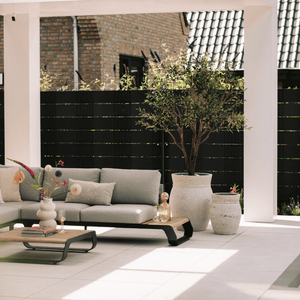 Kunstolijfboom 'Verdi' van 200 cm hoog, gepresenteerd onder een veranda. De kunstboom met zijn 3366 levensechte bladeren, realistische glasvezel stam, en houten takken voegt een elegante mediterrane charme toe aan de buitenruimte. De foto toont hoe de kunstolijfboom prachtig integreert in de veranda, waardoor een sfeervolle en luxe uitstraling ontstaat.