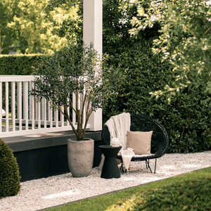 Kunstolijfboom 'Verdi' van 200 cm hoog, gepresenteerd in een luxe tuinsetting naast een zwarte rieten stoel. De foto toont de kunstboom met zijn 3366 levensechte bladeren en realistische glasvezel stam, die elegant samengaan met de stijlvolle zwarte rieten stoel en bijdragen aan een verfijnde mediterrane sfeer in de tuin.